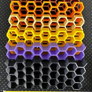 May include: A stack of six colorful honeycomb-patterned grids in pink, orange, yellow, gold, purple, and black. A yellow measuring tape is positioned at the base, showing the width of the grids is approximately 6 inches across the bottom edge.
