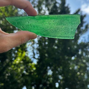 May include: A translucent, emerald green shard of glass with a textured surface and visible air bubbles. The piece is elongated and irregular in shape, featuring a straight edge on one side and a slightly curved, tapered edge on the other.