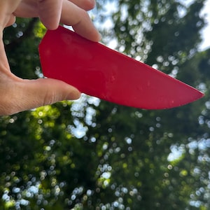 May include: A solid red, smooth, flat, wedge-shaped plastic tool with a pointed tip and a straight, thick edge. The object has a glossy finish and a simple, uniform design, appearing as a handheld implement for scraping or smoothing applications.