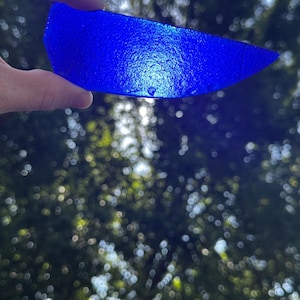 May include: A translucent, vibrant cobalt blue piece of sea glass with a textured, pebbled surface. The shard is shaped like a curved, elongated crescent with a pointed end, allowing light to pass through its deep blue, semi-opaque glass body.