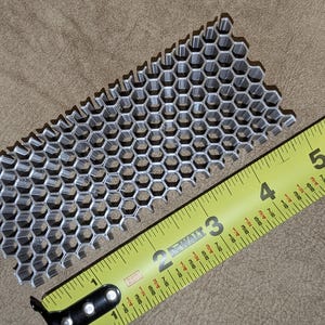 Op de afbeelding: Een zilverkleurig honingraatvormig metalen stuk met een gele rolmaat. Het honingraatstuk is ongeveer 15 cm lang. De rolmaat is geel met zwarte markeringen.