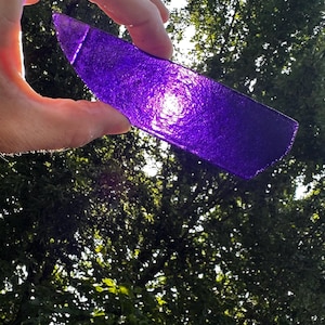 May include: A translucent, deep purple piece of textured glass with a slightly curved, elongated shape. The glass has a rough, uneven surface that catches the light, creating a vibrant, glowing effect when held up against the bright outdoor sky.
