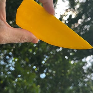 May include: A bright yellow, semi-translucent, flat, wedge-shaped plastic tool with a smooth surface and a pointed tip. The object is held against a bright, out-of-focus background of green foliage and dappled sunlight, highlighting its vibrant color.