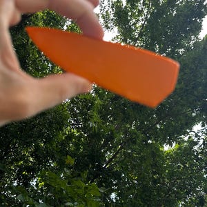 May include: A translucent, vibrant orange, wedge-shaped object with a smooth, flat surface. The piece features a pointed end and a wider, slightly angled base, displaying a uniform solid color throughout its structure when held up to the light.