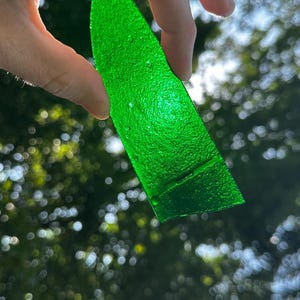 May include: A translucent, vibrant green glass shard with a textured, pebbled surface. The piece is elongated and tapered, resembling a leaf or blade shape, and allows light to pass through its semi-transparent, emerald-colored material.