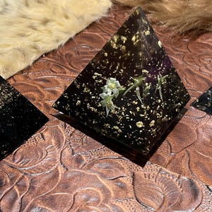 May include: Three black resin pyramids with gold leaf accents. The largest pyramid features small white flowers. The pyramids are displayed on a brown, textured surface, with a soft, furry background.