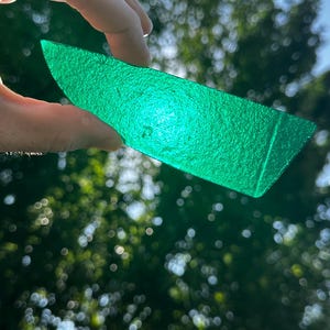 May include: A translucent, vibrant green glass shard with a textured, uneven surface. The piece is elongated and angular, catching the light to reveal a bright emerald hue throughout its semi-transparent body, showcasing a rough, organic finish.