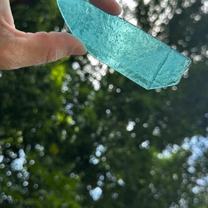 May include: A translucent, light turquoise-colored piece of sea glass with a curved top edge and a flat bottom edge. The glass has a textured, frosted surface that allows light to pass through, revealing a vibrant, cool-toned aqua hue.