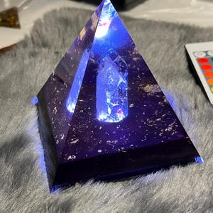 Puede incluir: Una pirámide de color púrpura oscuro con una luz azul brillante que emana desde el interior. Un cristal transparente está centrado dentro de la pirámide. La pirámide está sobre una superficie gris y esponjosa. Un mando a distancia es visible en el fondo.