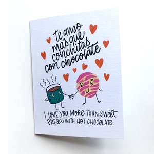 Op de afbeelding: Een kaart met een cartoonillustratie van een kopje warme chocolademelk en een zoet broodje met de tekst "Te amo mas que conchitas con chocolate" en "I love you more than sweet bread with hot chocolate".