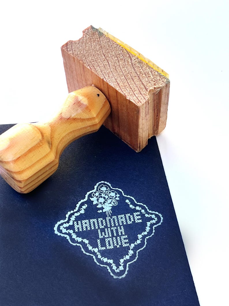 Puede incluir: Un sello de madera con mango se presiona sobre una superficie azul oscuro, revelando un dise&ntilde;o floral azul claro con las palabras "HANDMADE WITH LOVE". El sello est&aacute; hecho de madera clara.