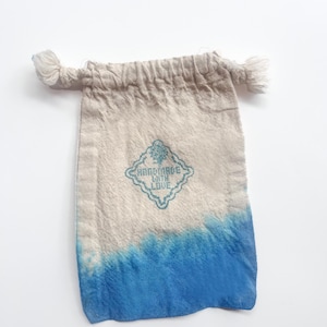 Puede incluir: Peque&ntilde;a bolsa de tela beige con efecto degradado azul en la parte inferior. La bolsa presenta el texto "HANDMADE WITH LOVE" en un dise&ntilde;o turquesa en forma de diamante. La parte superior de la bolsa tiene dos borlas anudadas.