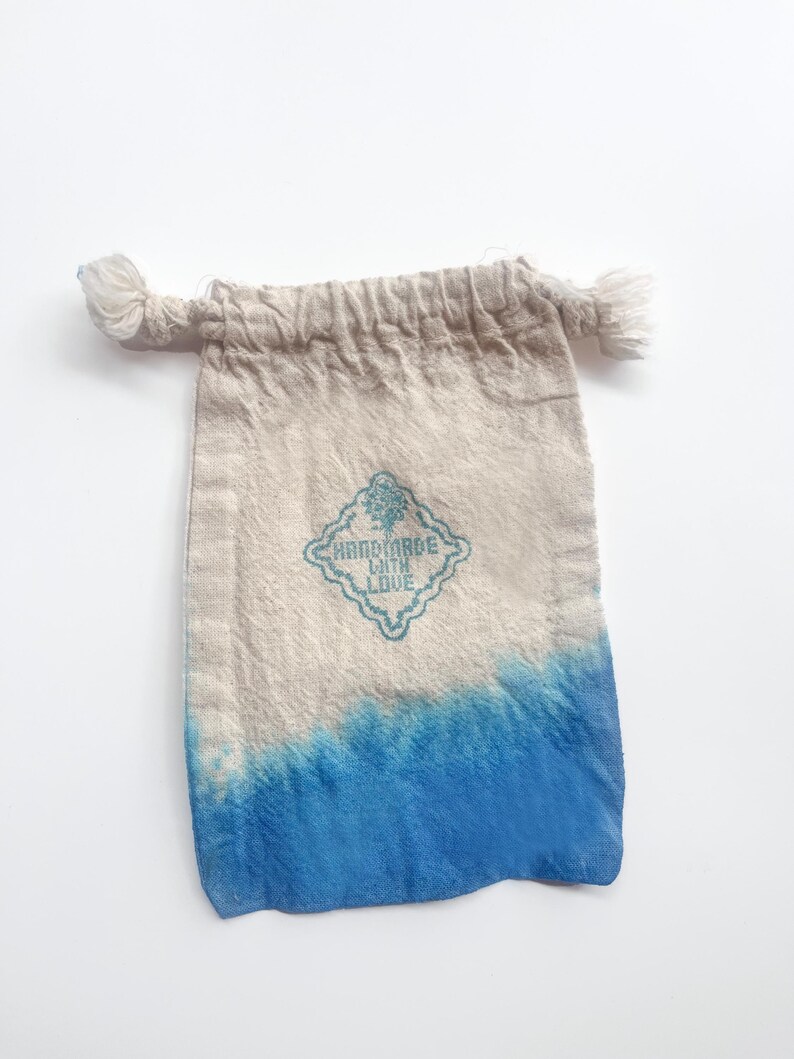 Puede incluir: Peque&ntilde;a bolsa de tela beige con efecto degradado azul en la parte inferior. La bolsa presenta el texto "HANDMADE WITH LOVE" en un dise&ntilde;o turquesa en forma de diamante. La parte superior de la bolsa tiene dos borlas anudadas.