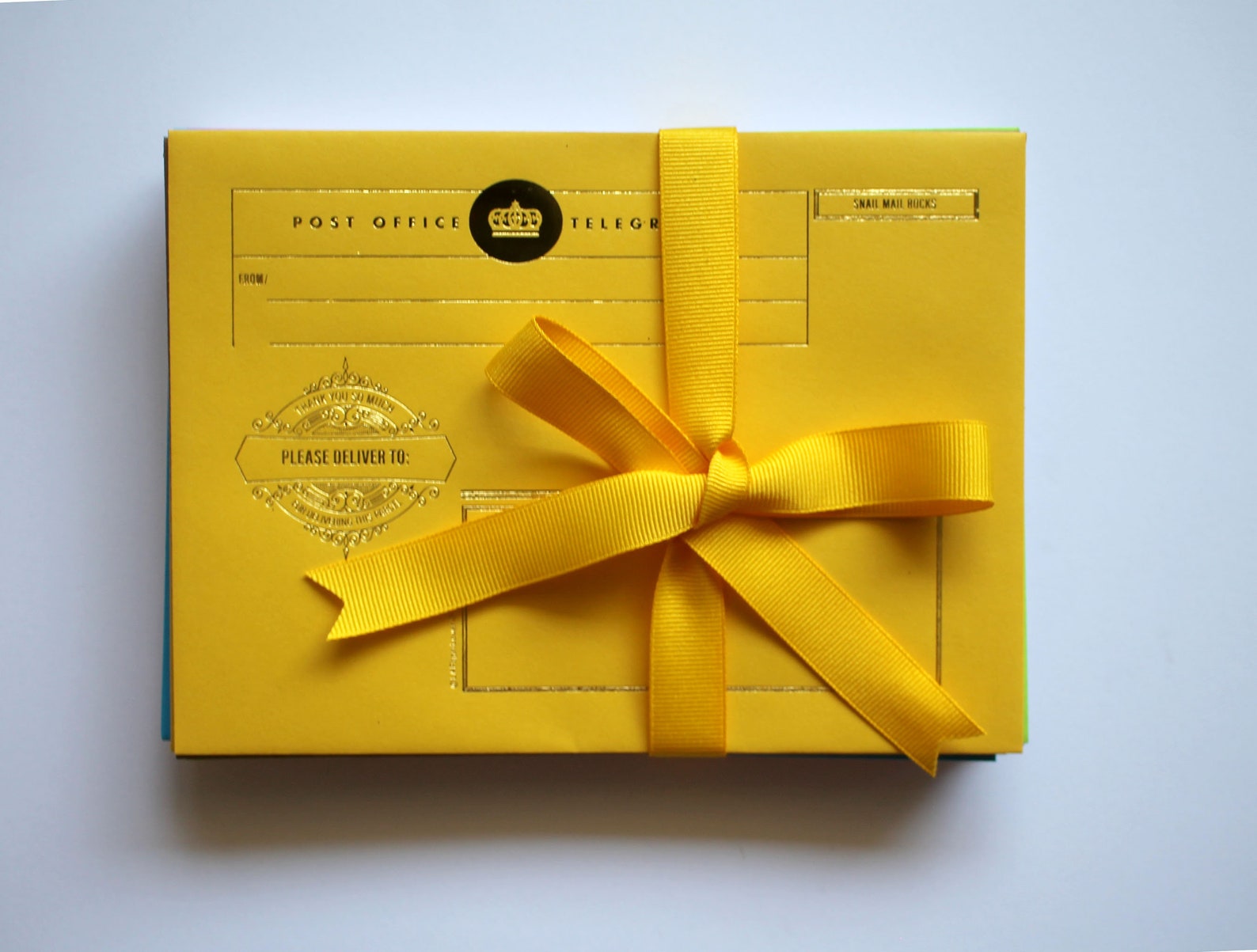 A yellow stationery set with a yellow bow and a golden crown emblem on a black circle. From left to right and up to down, the following in gold lettering: POST OFFICE, TELEGRAM, SNAIL MAIL ROCKS (in smaller lettering), and PLEASE DELIVER TO on top of a crest with the words THANK YOU SO MUCH (in smaller lettering)