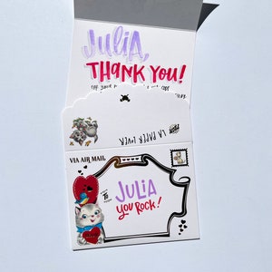 Puede incluir: Una tarjeta de felicitaci&oacute;n blanca con el nombre "Julia" en morado y las palabras "THANK YOU!" en rojo. La tarjeta presenta ilustraciones de gatos, corazones y un sello postal. El texto "JULIA you Rock!" tambi&eacute;n es visible.
