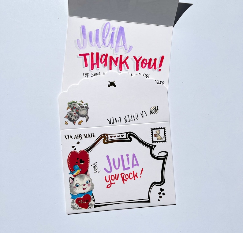 Puede incluir: Una tarjeta de felicitaci&oacute;n blanca con el nombre "Julia" en morado y las palabras "THANK YOU!" en rojo. La tarjeta presenta ilustraciones de gatos, corazones y un sello postal. El texto "JULIA you Rock!" tambi&eacute;n es visible.