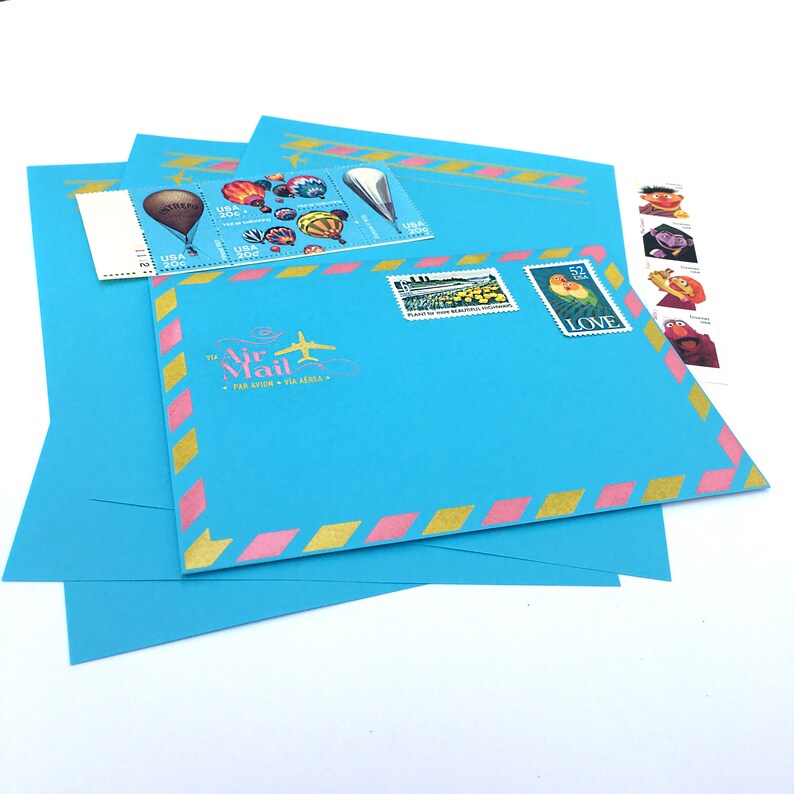 May include: A blue envelope with a pink and gold striped border. The envelope has a "Via Air Mail" stamp with a plane and the text "Par Avion - Via Aerea". There are several stamps on the envelope, including a stamp with a picture of a hot air balloon and a stamp with a picture of two birds.