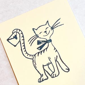 May include: A cream-colored card featuring a whimsical illustration of a smiling cat with a bow tie, holding a letter in its tail. The black ink drawing has a vintage, hand-drawn aesthetic, perfect for stationery or greeting cards.