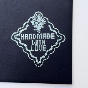 Puede incluir: Primer plano de una tarjeta azul oscuro con un dise&ntilde;o floral azul claro y las palabras "HANDMADE WITH LOVE" en un estilo de punto de cruz. El dise&ntilde;o est&aacute; enmarcado por un borde decorativo.