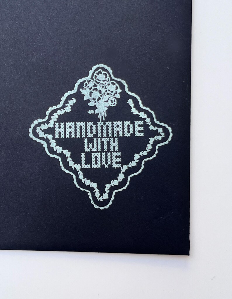 Puede incluir: Primer plano de una tarjeta azul oscuro con un dise&ntilde;o floral azul claro y las palabras "HANDMADE WITH LOVE" en un estilo de punto de cruz. El dise&ntilde;o est&aacute; enmarcado por un borde decorativo.