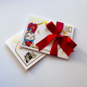 Puede incluir: Dos cajas de regalo blancas con detalles dorados y un lazo de sat&eacute;n rojo. Una caja presenta una ilustraci&oacute;n vintage de un gato sosteniendo un coraz&oacute;n con "HELLO!" y las palabras "VIA AIR MAIL". La otra caja tiene un dise&ntilde;o floral dorado.