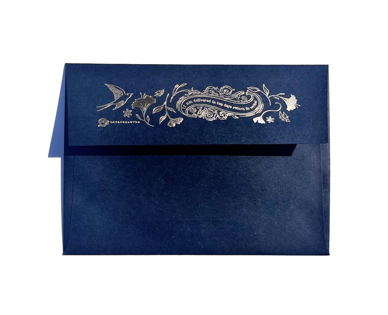 May include: A navy blue envelope with a silver foil design. The design includes a bird, floral elements, and text that reads "Not delivered in ten days return to sender" and "LAFAYETTE LOVES". The envelope is on a white background.