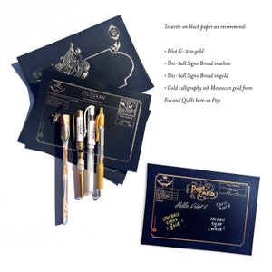 May include: A collection of black stationery items with gold accents, including postcards and telegrams. Several pens are arranged next to the stationery. The text on the postcard reads "Hello Pilot G" and "Uni-ball Signo - gold".