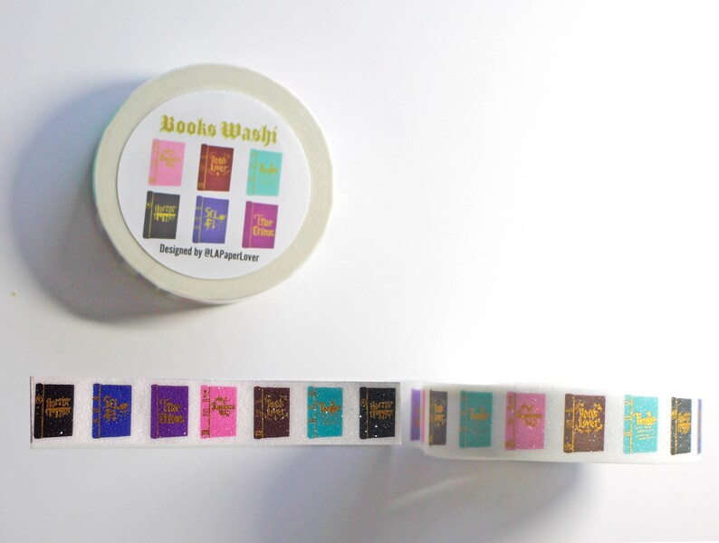 K&ouml;nnte beinhalten: Eine Rolle Washi-Tape mit goldenem Glitzer-Hintergrund und einem sich wiederholenden Muster aus goldenen Buchumschl&auml;gen mit Titeln wie "The Crime" und "Sea of Fi". Das Band ist mit "Booko Washi" und "Designed by @LAPaperLover" beschriftet.