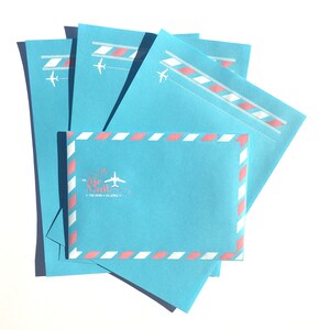 May include: Stack of blue airmail envelopes with pink and white stripes and a plane graphic. The envelope has the text "Via Air Mail" with the tagline "Par Avion - Via Aerea".