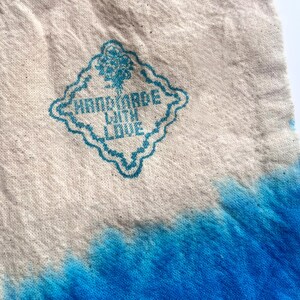 Puede incluir: Primer plano de un art&iacute;culo de tela con un dise&ntilde;o te&ntilde;ido anudado azul. La parte superior es de color beige natural, con un emblema azul en forma de diamante que dice "HANDMADE WITH LOVE". La parte inferior de la tela es azul vibrante.