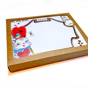 Puede incluir: Una caja de papel kraft con una ventana decorativa, que contiene art&iacute;culos de papeler&iacute;a. La papeler&iacute;a presenta una ilustraci&oacute;n vintage de un gato sosteniendo un coraz&oacute;n que dice "Hello". El dise&ntilde;o incluye un coraz&oacute;n rojo, un p&aacute;jaro azul y detalles dorados. Tambi&eacute;n se ven las palabras "To" y "Air Mail".