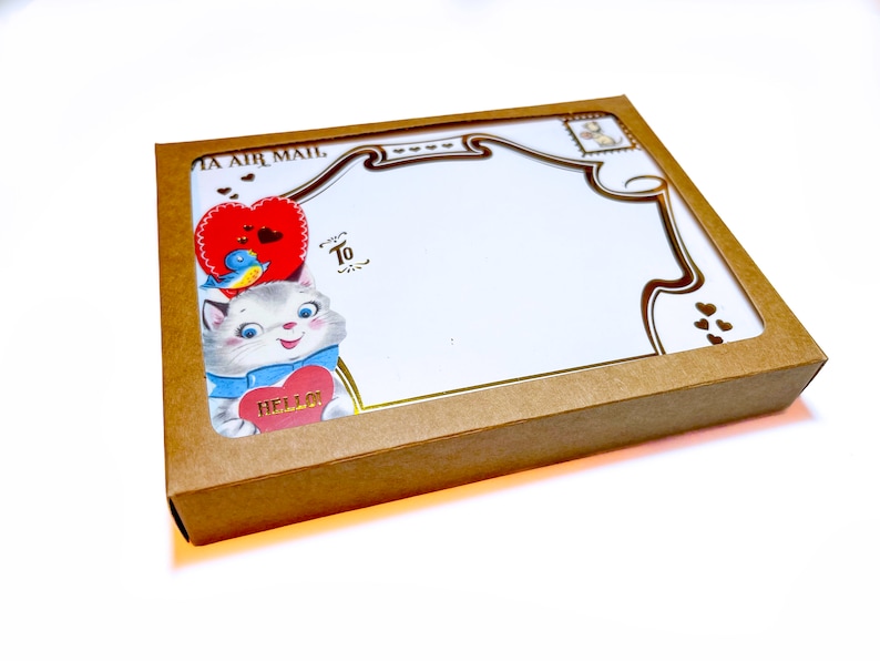 Puede incluir: Una caja de papel kraft con una ventana decorativa, que contiene art&iacute;culos de papeler&iacute;a. La papeler&iacute;a presenta una ilustraci&oacute;n vintage de un gato sosteniendo un coraz&oacute;n que dice "Hello". El dise&ntilde;o incluye un coraz&oacute;n rojo, un p&aacute;jaro azul y detalles dorados. Tambi&eacute;n se ven las palabras "To" y "Air Mail".