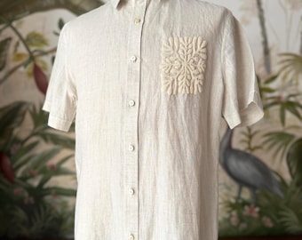 May include: A short-sleeved, button-down shirt in a light beige color. The shirt features a decorative, embroidered design on the chest pocket. The shirt is made of a textured fabric and has a classic collar.