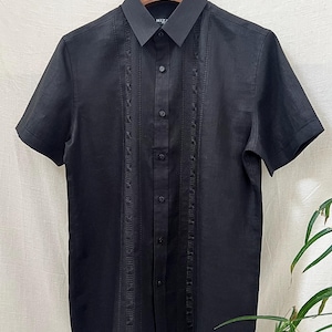Puede incluir: Camisa negra de manga corta con cuello clásico y cierre de botones. La camisa presenta intrincados paneles verticales bordados a ambos lados de la tapeta de botones. La marca "MIZ" es visible en el cuello.