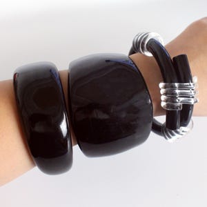 Peut inclure: Trois bracelets noirs sont présentés sur un poignet. Deux sont de larges bracelets brillants. Le troisième bracelet est noir avec des accents métalliques argentés. Les bracelets sont un mélange de styles modernes et classiques.