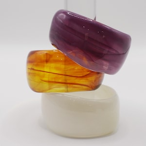 Puede incluir: Tres brazaletes gruesos apilados en tonos morado, ámbar y crema. El brazalete morado tiene un efecto veteado, el ámbar tiene un tono dorado y el brazalete crema es opaco. Estas pulseras de resina son un accesorio de moda audaz.