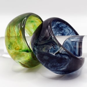 May include: Two resin bangles. One is green with yellow swirls, the other is dark blue with white swirls. The bangles are thick and rounded, with a glossy finish. They are displayed on a clear, cylindrical support.
