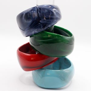 Op de afbeelding: Vier gestapelde, dikke armbanden in tinten blauw, groen en rood. De bovenste armband is donkerblauw met witte marmering. De tweede is glanzend groen, de derde is felrood en de onderste is lichtblauw.