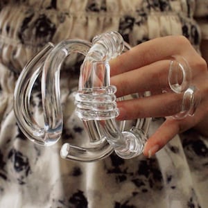 Könnte beinhalten: Armband und Ringe aus klarem Glas. Das Armband hat ein geschlungenes Design mit einer gewickelten Mitte. Zwei klare Glasringe befinden sich an den Fingern. Der Schmuck ist transparent und scheint aus Glas zu sein.