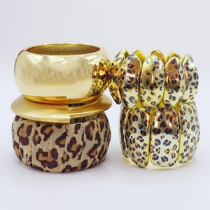 May include: Three gold and leopard print bracelets. One is a wide, hammered gold bangle. Another is a gold band with a leopard print fabric. The third is a stretch bracelet with gold and leopard print segments.