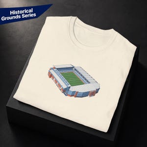 Può includere: Una t-shirt color crema con un'illustrazione grafica di uno stadio con un campo verde e tribune blu e bianche. Il testo Historical Grounds Series è stampato in bianco su uno sfondo triangolare blu scuro nell'angolo in alto a sinistra.