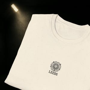 Puede incluir: Camiseta crema de cuello redondo con un pequeño diseño floral y la palabra "LEEDS" impresa debajo en negro. La camiseta es de tela moteada.