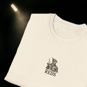 May include: Cream-colored t-shirt featuring a graphic of a man with a bow and arrow, with the word "REDS" printed below. The shirt has a crew neck and a speckled texture.