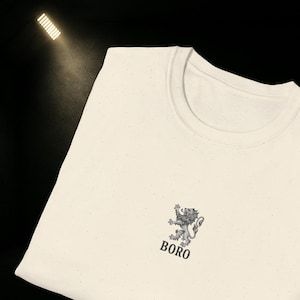 May include: Cream-colored t-shirt with a crew neck. The shirt features a gray lion crest design with the word "BORO" printed below it. The shirt is made of a speckled fabric.