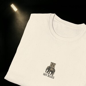 May include: Off-white t-shirt with a crew neck and a graphic of an elephant carrying a building on its back. The text "SKY BLUES" is below the graphic. The shirt is made of a speckled fabric.