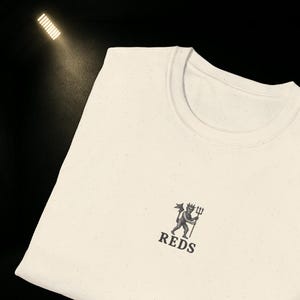 May include: Cream-coloured crew neck t-shirt with a small graphic of a devil holding a trident, and the word "REDS" printed below. The shirt has a speckled texture.