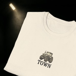 May include: Cream-colored t-shirt featuring a detailed illustration of a tractor. The tractor is in shades of gray and black, with the word "TOWN" printed below it in a bold, black font. The shirt has a classic crew neck design.