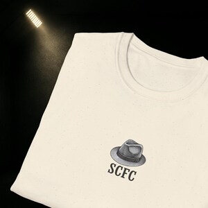 Könnte beinhalten: Cremefarbenes T-Shirt mit einer kleinen Grafik eines Fedora-Huts und den Buchstaben "SCFC" darunter. Das Shirt hat einen Rundhalsausschnitt und eine gesprenkelte Textur.