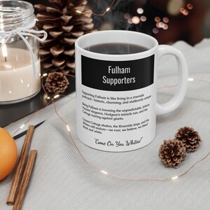 May include: White ceramic mug with the text "Fulham Supporters" in a black rectangle. The mug has a curved handle and is filled with a dark liquid. Additional text includes team slogans and phrases.