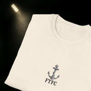 Könnte beinhalten: Cremefarbenes T-Shirt mit einer grauen Ankergrafik und den Buchstaben "FTFC" darunter. Das Shirt hat einen Rundhalsausschnitt und eine gesprenkelte Textur. Das Bild wird von der oberen linken Ecke beleuchtet.